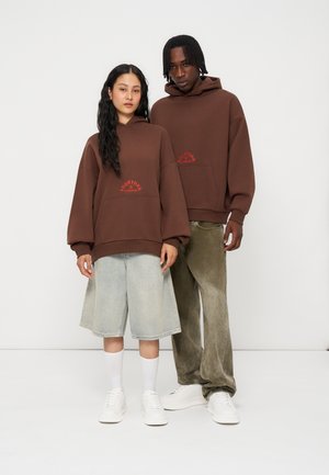 To personer iført matchende brune hættetrøjer med rød tekst, den ene i lyse denimshorts og hvide sko, den anden i løse olivengrønne bukser og hvide sko.