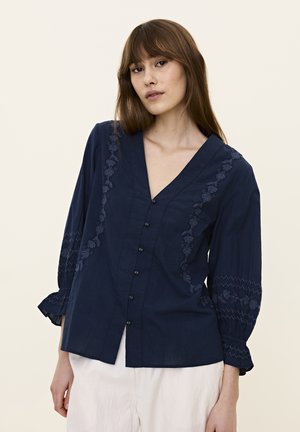 Blusa blu navy con profondo scollo a V, bottoni frontali e delicata ricamo floreale lungo le cuciture e le maniche, con polsini elasticizzati.