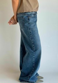 Niebieskie dżinsowe spodnie o szerokich nogawkach i wysokim stanie, z widocznym przeszyciem oraz dwoma bocznymi kieszeniami, w połączeniu z oliwkowymi półbutami z ostrym noskiem.