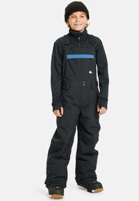 Sorte vinteroveraller med en blå vandret stripe, justerbare stropper og en lynlåslomme; parret med en sort langærmet top og en hue.