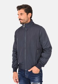 Bomber jacket di un blu navy scuro con colletto alto, zip frontale e tasche laterali. Presenta polsini e orlo a coste, realizzato in materiale leggero.