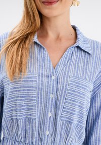 Blau gestreiftes Hemd mit Kragen, zwei Fronttaschen und Knopfverschluss. Der Stoff ist strukturiert und weist abwechselnde dicke und dünne Streifen auf.