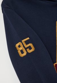 Marine gebreide mouw met gouden geborduurd nummer "85." De stof heeft een gestructureerde afwerking met een onderscheidend stikseldetail.