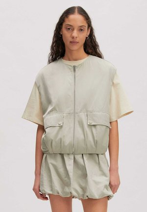 Vrouw met lang krullend haar draagt een lichtgroen vest met ritssluiting en klepzakken over een beige top met korte mouwen en bijpassende losse short.