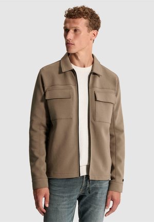 Homme aux cheveux bouclés portant une veste beige zippée par-dessus un pull blanc et un jean bleu, regardant vers la gauche sur un fond uni.