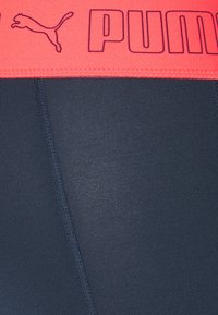Leggings aderenti blu navy con un ampio elastico fucsia neon che presenta un logo Puma in nero. Il tessuto è liscio con una leggera lucentezza e elasticità.