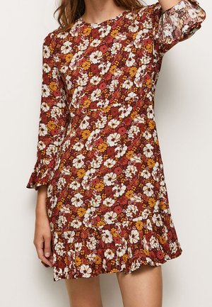 Femme portant une robe courte à manches longues avec un motif floral rouge, blanc et jaune et un ourlet volanté.