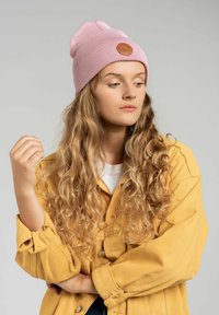 Różowa dziana czapka z brązową skórzaną łatą, w zestawieniu z żółtą koszulą zapinaną na guziki. Modelka ma falujące, długie włosy i pozuje w zamyśleniu.