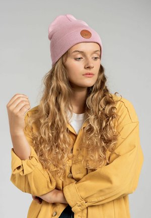 Różowa dziana czapka z brązową skórzaną łatą, w zestawieniu z żółtą koszulą zapinaną na guziki. Modelka ma falujące, długie włosy i pozuje w zamyśleniu.