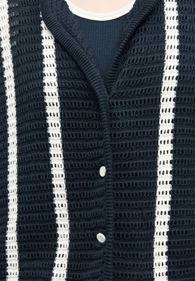 Μαύρο ζακέτα με ανοιχτό πλέξιμο, κάθετες λευκές ρίγες και λευκά κουμπιά, φορεμένη πάνω από μαύρο πουκάμισο με ριγέ ύφασμα.