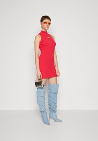 Mini abito rosso a coste con collo alto, abbinato a stivali slouchy azzurri fino alla coscia. La modella tiene in mano una piccola borsa nera e argentata.