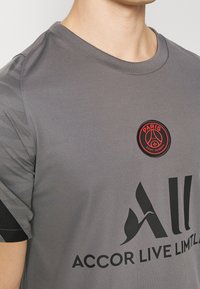 Grå träningsskjorta med korta ärmar, har rund halsringning, svart kant och en rund röd Paris Saint-Germain-logotyp på bröstet.