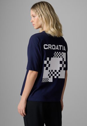Blondynka w granatowej koszuli z napisem "CROATIA" i pikselowym wzorem szachownicy na plecach, stojąca na szarym tle.