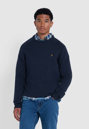 Marineblauwe gebreide sweatshirt met geribde manchetten, gedragen over een blauw-wit geruite blouse. Heeft een klein geel logo op de borst.
