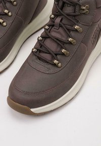 Bota de senderismo de cuero marrón con cordones de textil, ojales de metal, suela acolchada y una suela de goma elevada. Presenta un logo en el lateral.