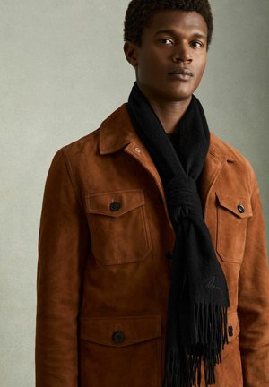 Jonge man die een bruine suède jas draagt met zakken met knopen en een zwarte sjaal met franjes om zijn nek geknoopt.