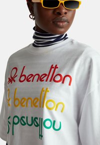 Bílé bavlněné tričko s vícero barevným potiskem v červené, žluté a zelené barvě. Nošeno s námořnicky modrým a bílým pruhovaným rolákem a žlutými slunečními brýlemi.