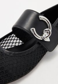 Zapato plano negro que presenta un upper de malla texturizada, una correa de cuero suave y un detalle de hebilla circular en tono plateado.