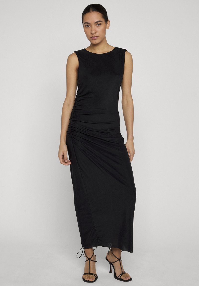 Femme portant une robe maxi noire sans manches à fronces et des talons noirs à brides, debout devant un fond clair uni.