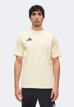 Giovane uomo in piedi di fronte che indossa una t-shirt Adidas beige chiaro e pantaloni Adidas neri su uno sfondo neutro.