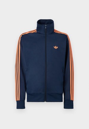 Temno modra tekaška jakna s polno zadrgo spredaj, visokim ovratnikom, oranžnimi črtami na rokavih in majhnim oranžnim logotipom na prsih.