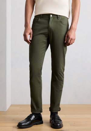 Man draagt olijfgroene opgerolde broek, beige mouwloos topje, zwarte sokken en zwarte leren loafers en staat op een houten vloer.