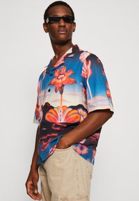 Cămașă cu mânecă scurtă, cu nasturi, având un imprimeu floral tropical în culori vibrante, în principal albastru, roșu și roz, cu un croi lejer.