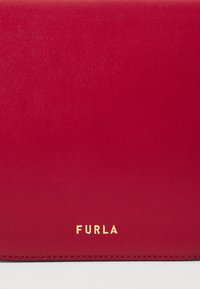 Portafoglio in pelle rossa con una superficie liscia, caratterizzato da un logo dorato "FURLA" e una cucitura ordinata lungo i bordi. Design rettangolare compatto.