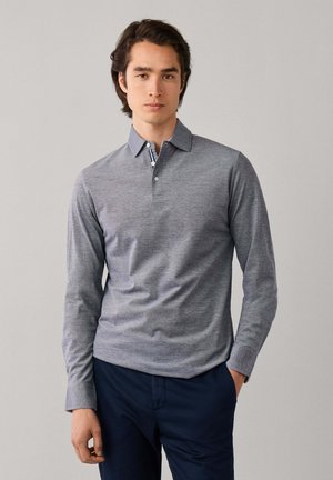 Jonge man met een grijs poloshirt met lange mouwen en donkere broek, staand tegen een effen lichte achtergrond.