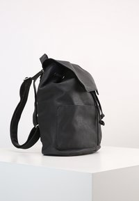 Sac à dos en cuir noir avec une fermeture à cordon, une poche frontale plate et une seule sangle ajustable. Texture lisse et forme arrondie.