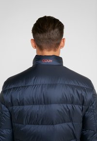 Marineblå dunjakke med quiltet mønster, høj krave og rødt logo bagpå. Materialet ser skinnende og glat ud.
