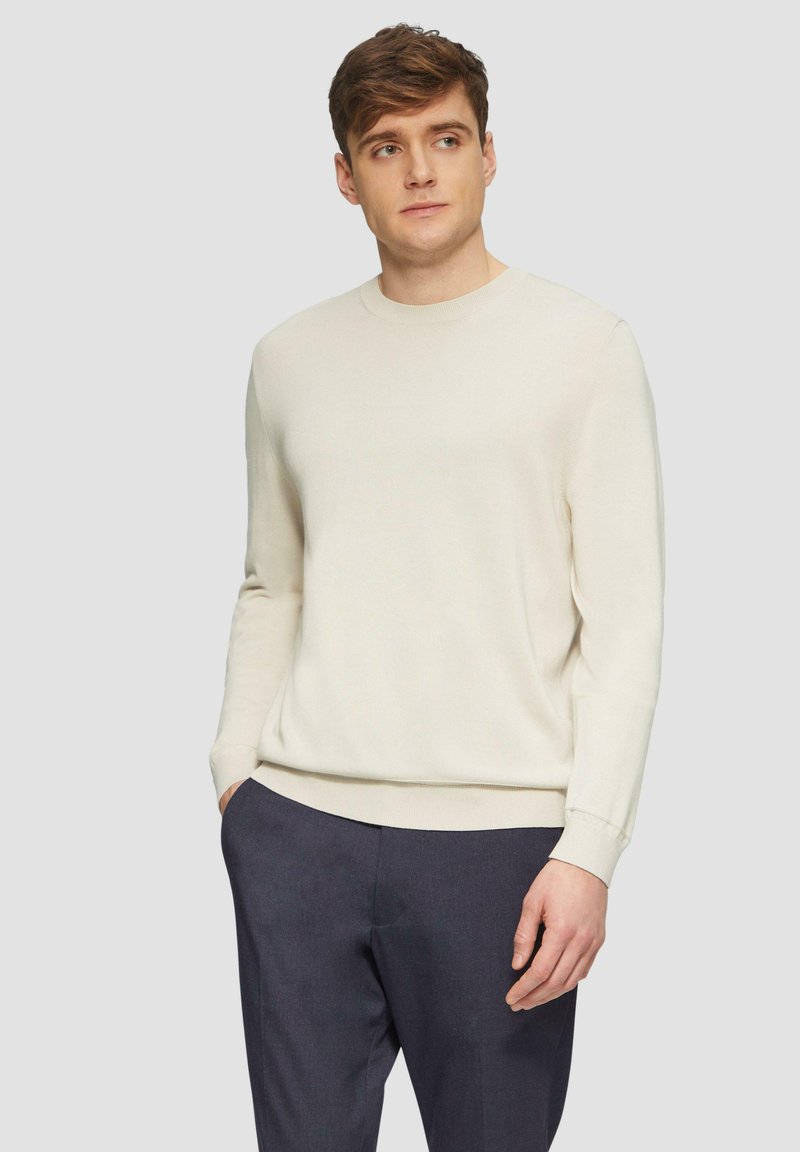 Pull à col rond beige clair avec des manches longues, en texture douce. Associé à un pantalon sombre, se tenant devant un fond gris clair.