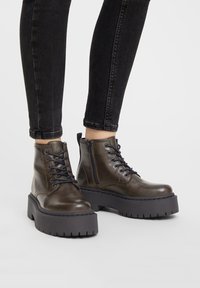 Bottines en cuir marron avec semelles compensées noires épaisses, laçage à l'avant et fermeture éclair sur le côté. Texture lisse avec des détails de couture contrastés.