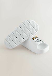 Sabots blancs à bout ouvert et semelle texturée. Présentent un design graphique sur la bride avec des lettres en gras et des accents de motifs.