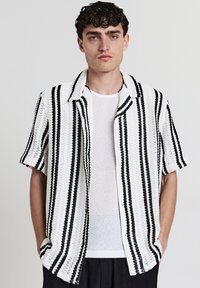 Giovane uomo con capelli ricci che indossa una camicia bianca a maniche corte con strisce verticali nere e una maglietta bianca trasparente sotto.