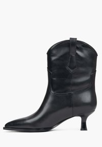 Czarny skórzany botek do kostki z ostrym czubkiem, zakrzywioną górą i krótkim obcasem w stylu szpilki. Posiada minimalne przeszycia i zaokrągloną pętelkę do ściągania.