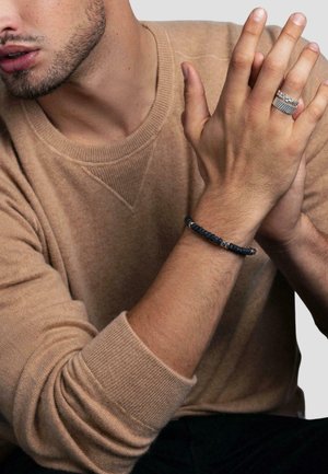 Perlekæde med mørkeblå strukturerede perler og sølvfarvede accenter, båret på et håndled. Model i en sandfarvet sweater med ribbet manchet.