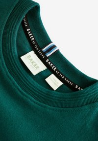 Tmavozelený sveter s okrúhlym výstrihom. Má textúrovanú látku, rebrovaný golier a tkané etikety v čiernej farbe a pruhovanej modrej.