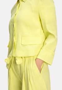 Linen two-piece outfit in bright yellow, featuring a collared jacket with button closure and pockets, paired with loose, pleated trousers.
