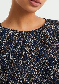 Haut bleu marine avec un col rond, présentant un tissu texturé avec un motif tacheté en marron, blanc et noir. Les détails incluent des plis.
