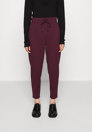 Osoba ubrana w bordowe spodnie z wysokim stanem i zwężanymi nogawkami z sznurkiem, czarną koszulę z długim rękawem oraz czarne mokasyny, stojąca na szarej podłodze.