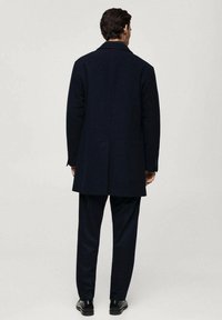 Cappotto di lana navy con colletto classico, apertura centrale sul retro e polsini con bottone, abbinato a pantaloni scuri e scarpe nere.