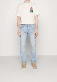 T-shirt di cotone beige con una grafica verde e arancione sulla tasca del petto, abbinata a jeans di denim blu chiaro e sneaker color sabbia.