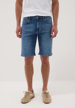 Mann trägt mittelblaue Jeansshorts, weißes T-Shirt und beige geschnürte Freizeitschuhe, steht vor einem schlichten hellen Hintergrund.