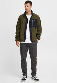 Olive green fleece jacket with a navy button-up front, chest pocket, and a smooth texture, paired with gray cargo pants and black sneakers.