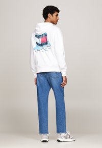 Felpa bianca con un grafico colorato sul retro, caratterizzato da un liquido blu e un logo, abbinata a jeans azzurri e sneakers grigie.
