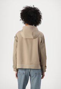 Giacca beige con cappuccio e vestibilità rilassata, presenta una texture liscia e maniche arrotolate. Indossata con jeans di denim blu chiaro.