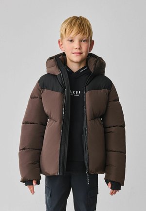 Braune Pufferjacke mit einer schwarzen Kapuze und Ärmeln, ausgestattet mit einem Reißverschluss und strukturierten Paneelen. Getragen über einem schwarzen Hoodie.