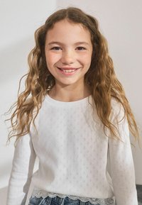 Camicia bianca a maniche lunghe con un motivo di occhiello texturizzato e orlo in pizzo a zigzag. Abbinata a pantaloni shorts in denim. Ragazza giovane con capelli ondulati che sorride.