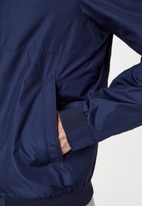 Marineblauwe bomberjack met een gladde, glanzende textuur. Voorzien van een voorzak en geribbelde manchetten. Gemaakt van lichtgewicht, waterafstotende stof.